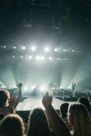 Concert crowd silhouettes with raised hands against bright stage lights creating beams through atmospheric haze.