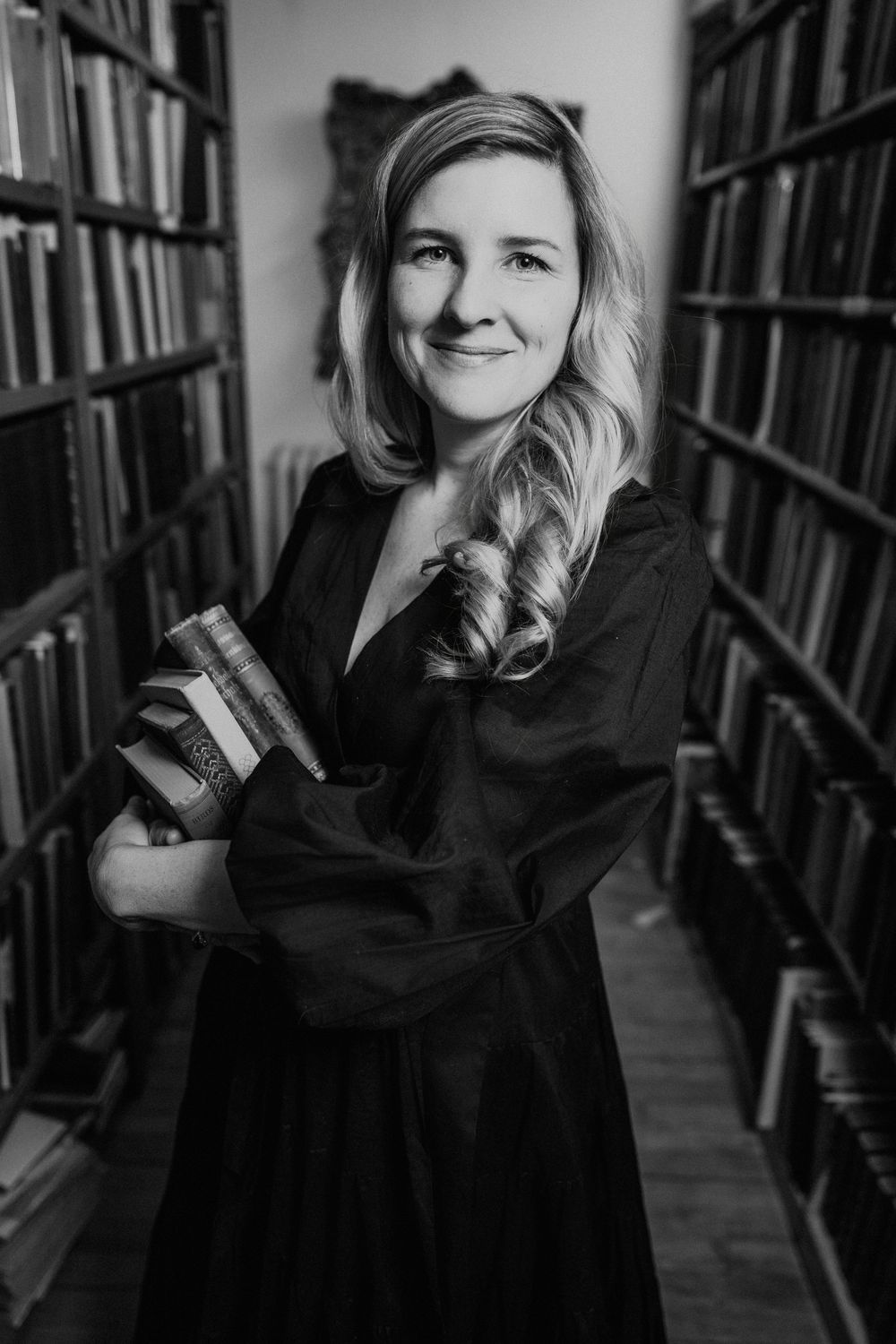 Black and white photo of a smiling woman holding books in a library aisle, wearing a dark dress.