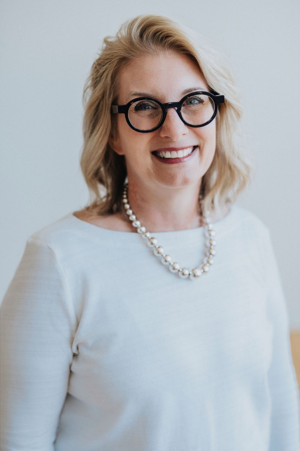 Smiling blonde woman wearing black round glasses, pearl necklace, and white sweater poses against a light background.
