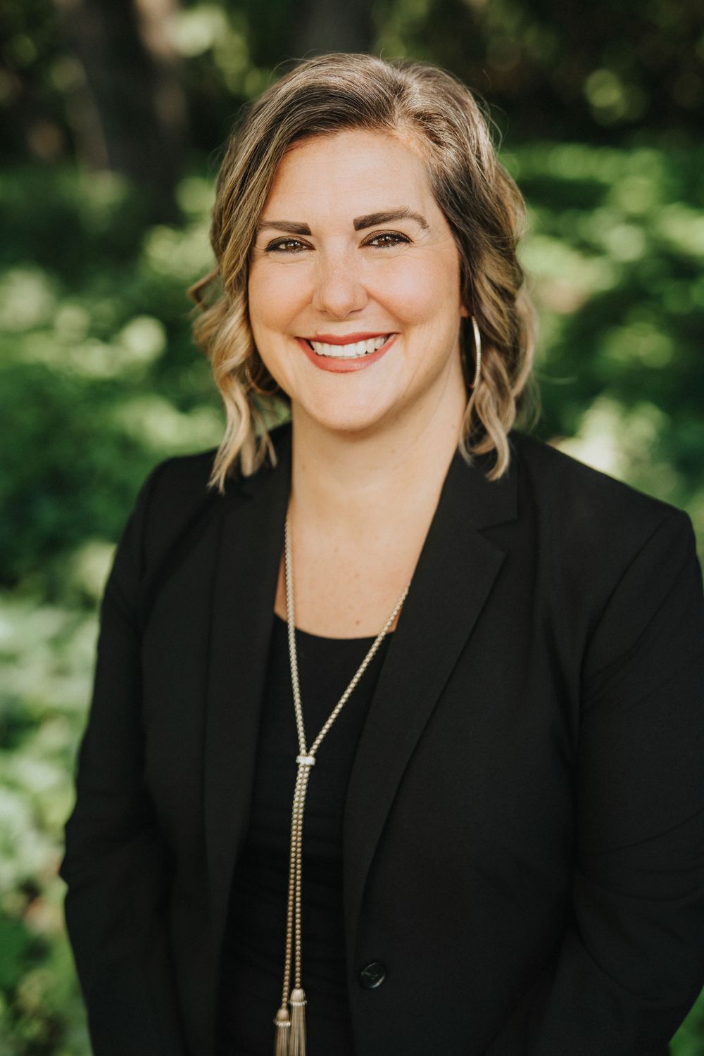 Professional woman in black blazer and long necklace smiling outdoors with lush green trees in background.