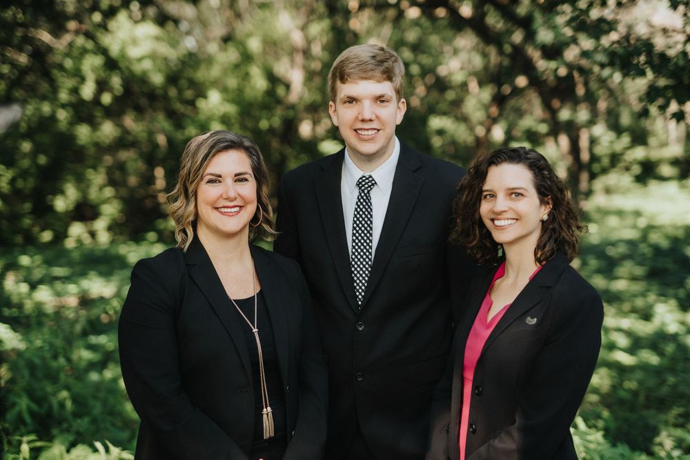 Three professionals in formal attire smiling outdoors, two women and one young man in a suit and tie.