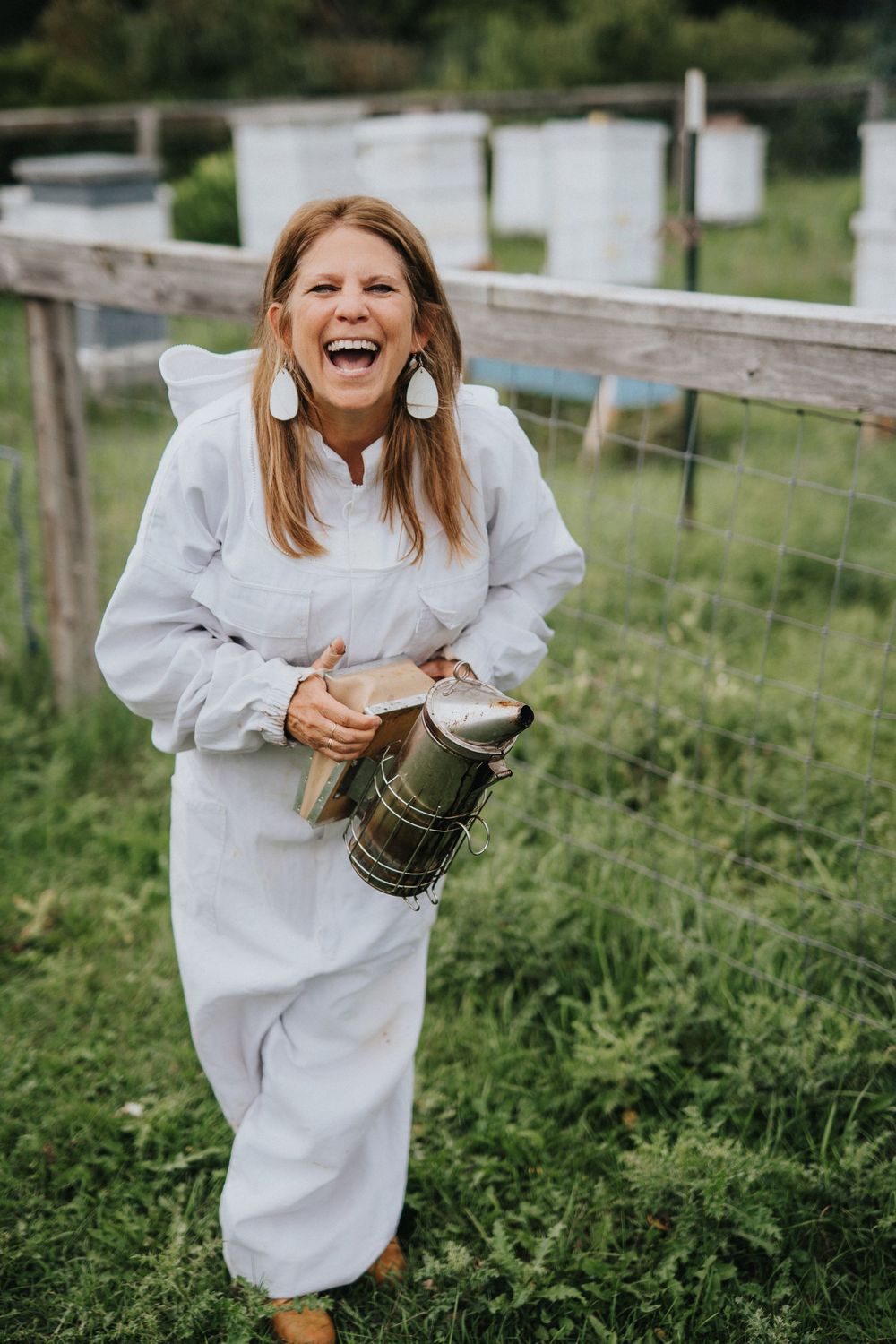 Smiling woman in white beekeeper suit holding a smoker tool near beehives in a green field.
