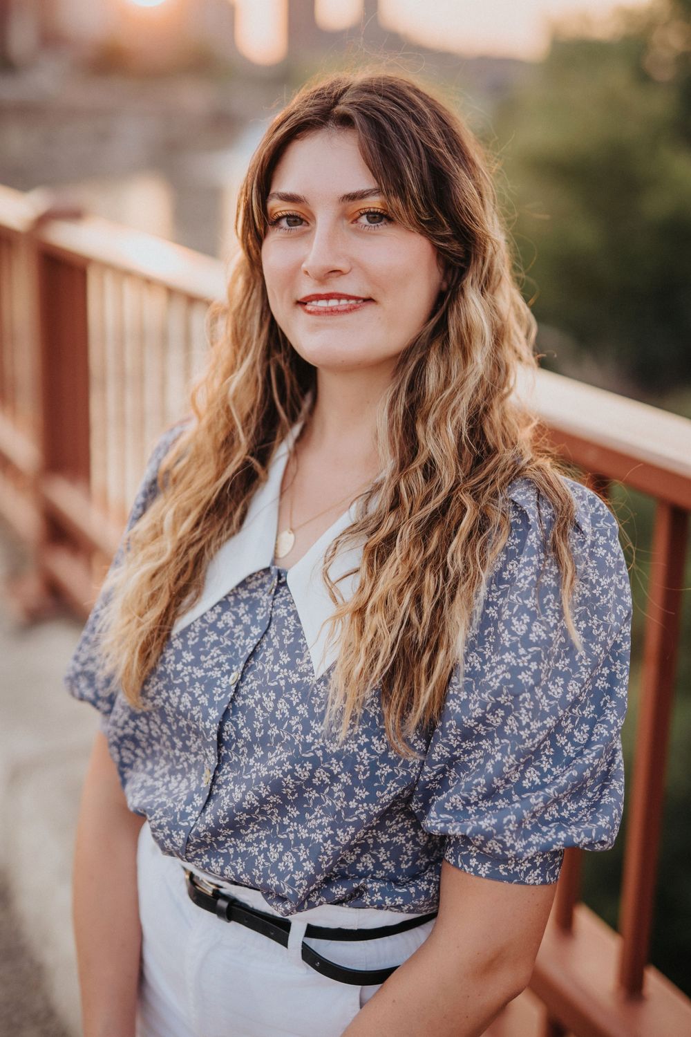 Young woman with long wavy hair smiling in a blue floral blouse on a wooden bridge at golden hour.