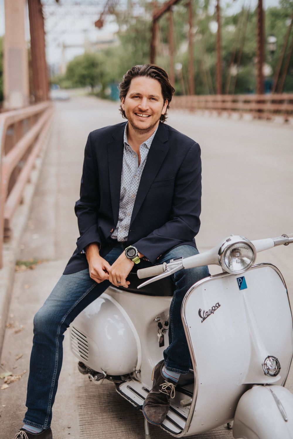 Smiling man in navy blazer sitting on a vintage white Vespa scooter on a bridge, casual professional style.