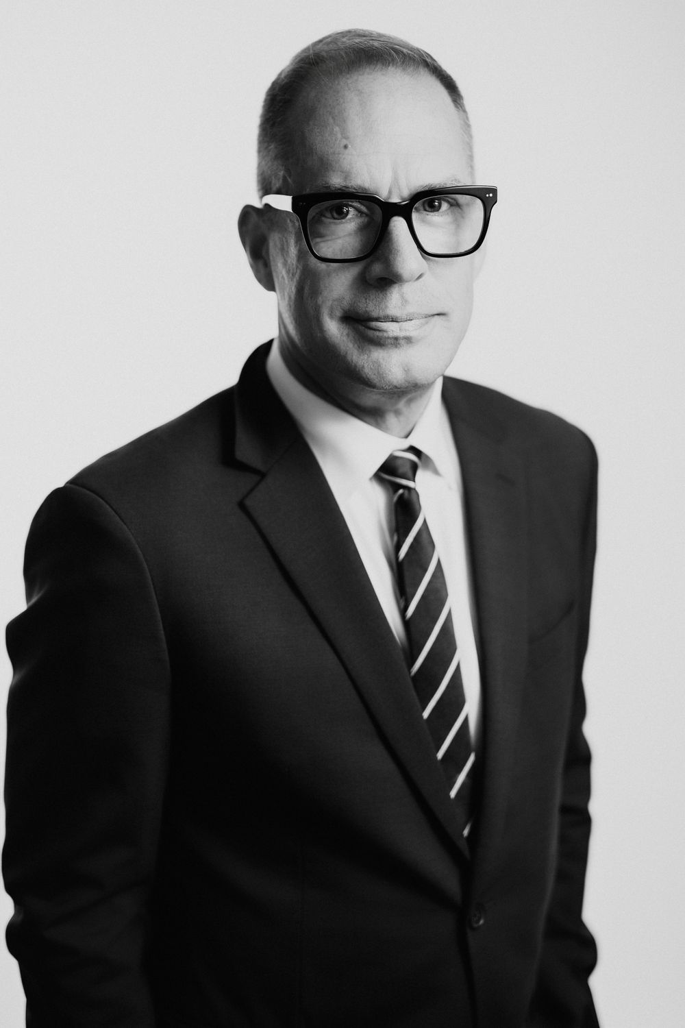 Black and white professional headshot of a middle-aged man in a suit and tie wearing bold-framed glasses.