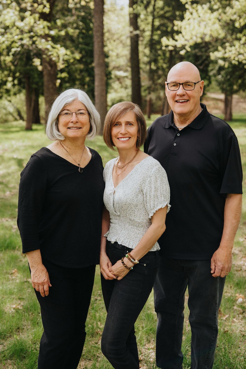 Three smiling adults pose outdoors in a lush green park setting, wearing casual black and white outfits.