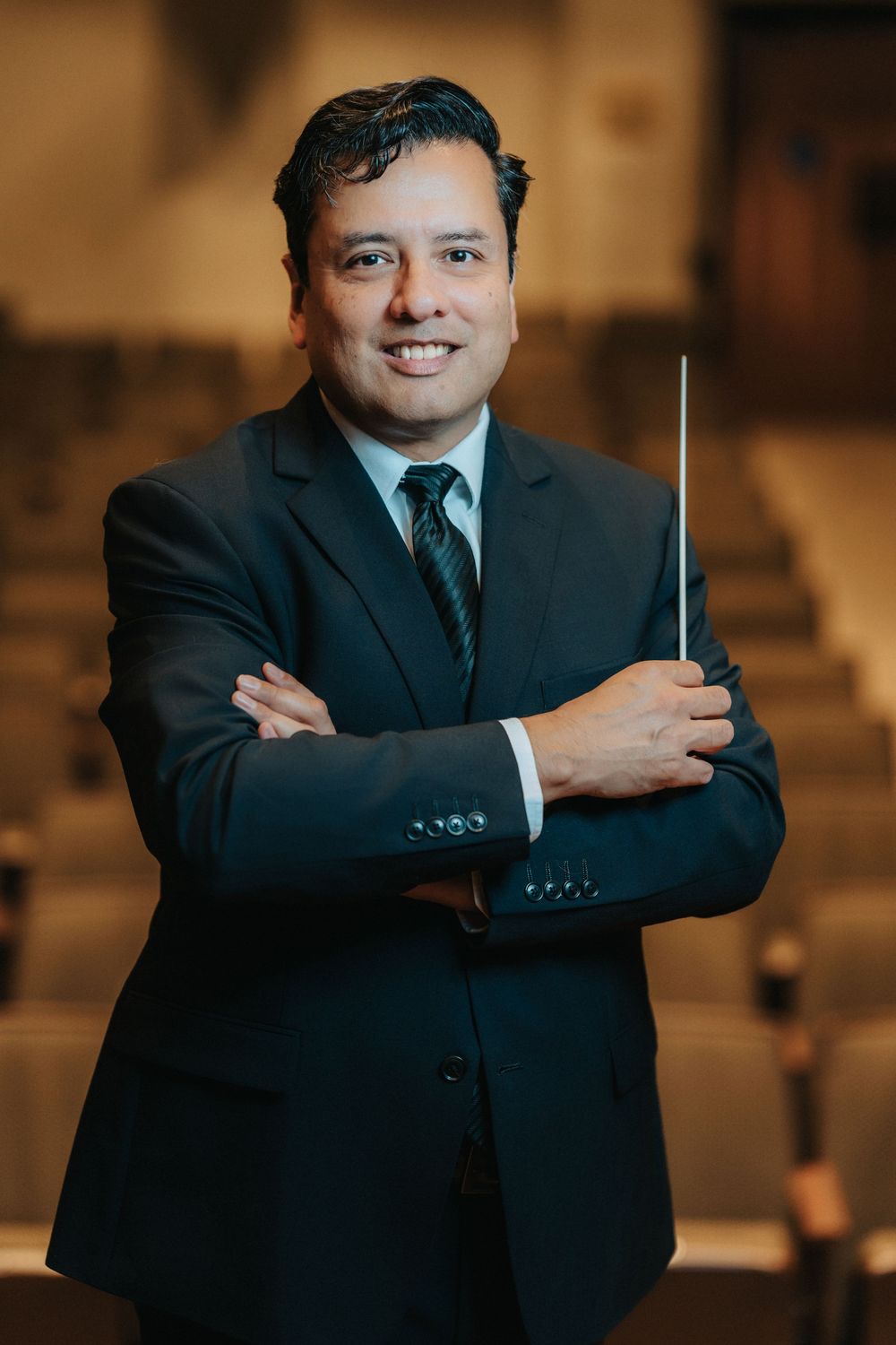 A smiling orchestra conductor in a dark suit holds a baton with arms crossed in an elegant concert hall.
