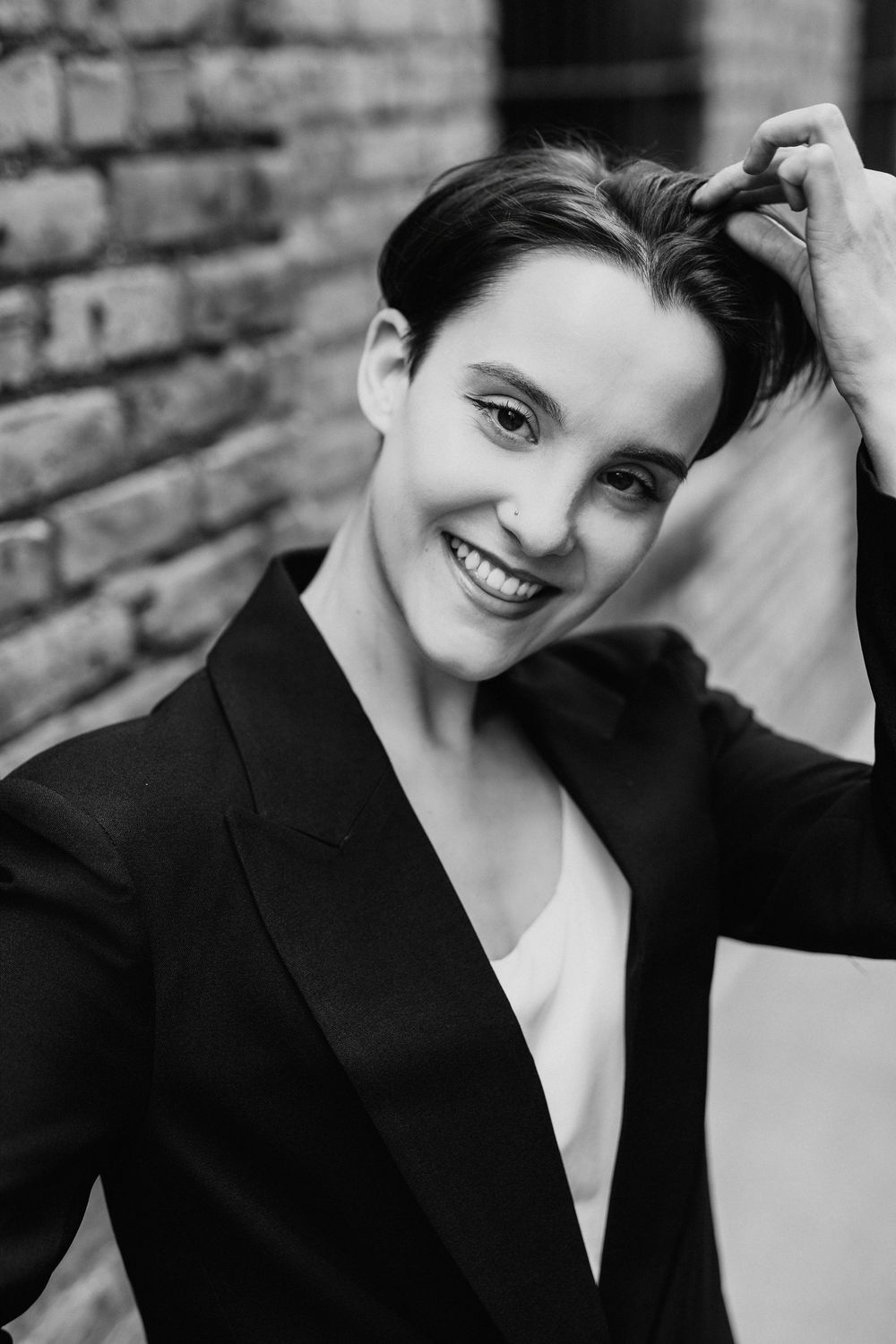 Black and white portrait of a smiling woman with short hair in a blazer, posing against a brick wall.