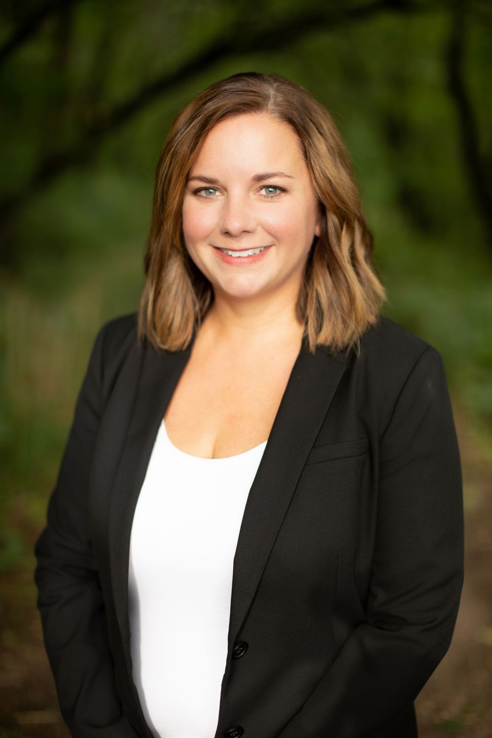 Professional headshot of a smiling woman in a black blazer and white top, posed outdoors with green foliage background.