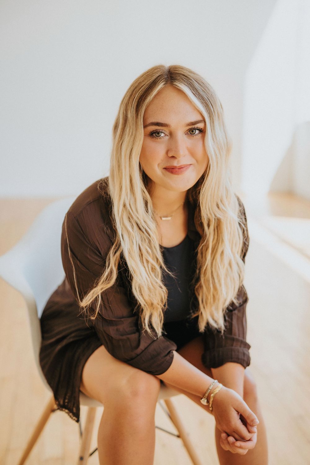 Young blonde woman in dark shirt sits on stool, smiling warmly in bright modern space.