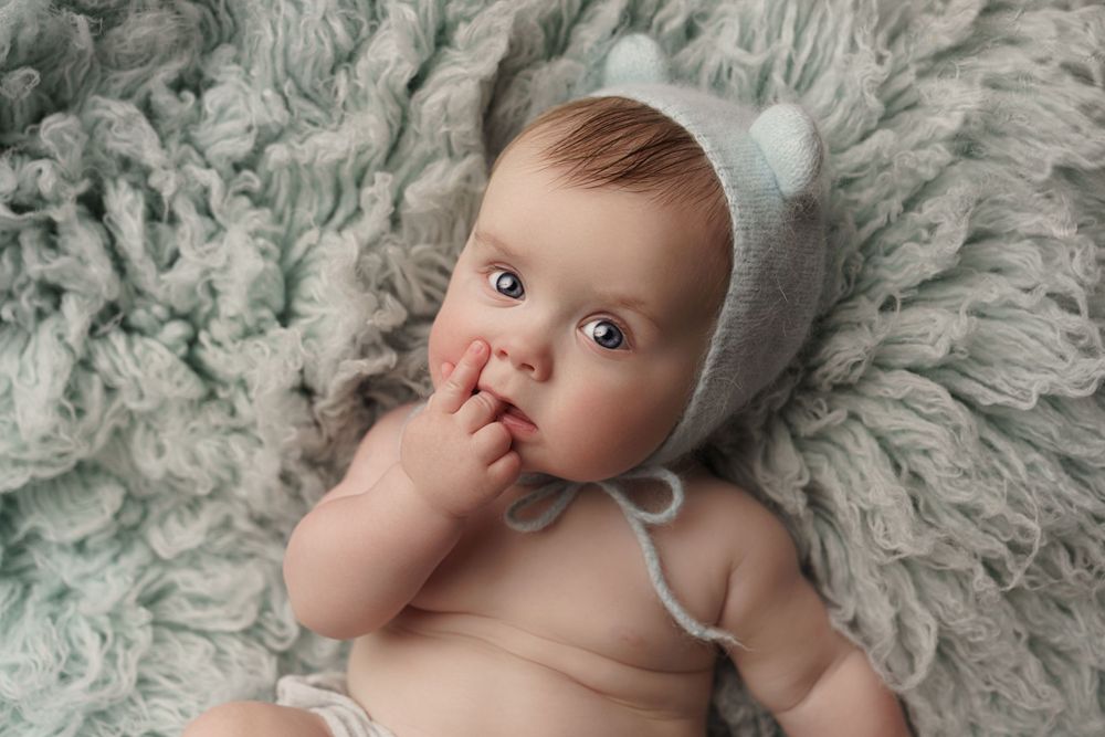 Baby lying on plush gray rug wearing white bonnet while sucking thumb.