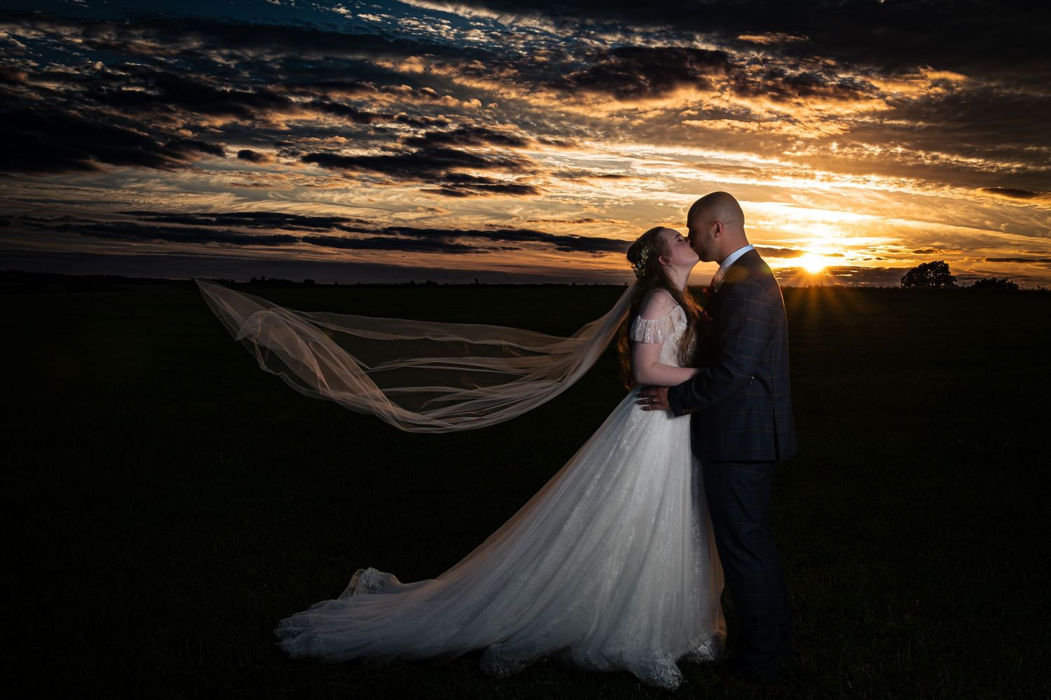 Bride and groom pose in the sunset against a dramatic sky.  Blue-gold skyscape.  Veil flowing out behind. Donnington park Farmhouse.