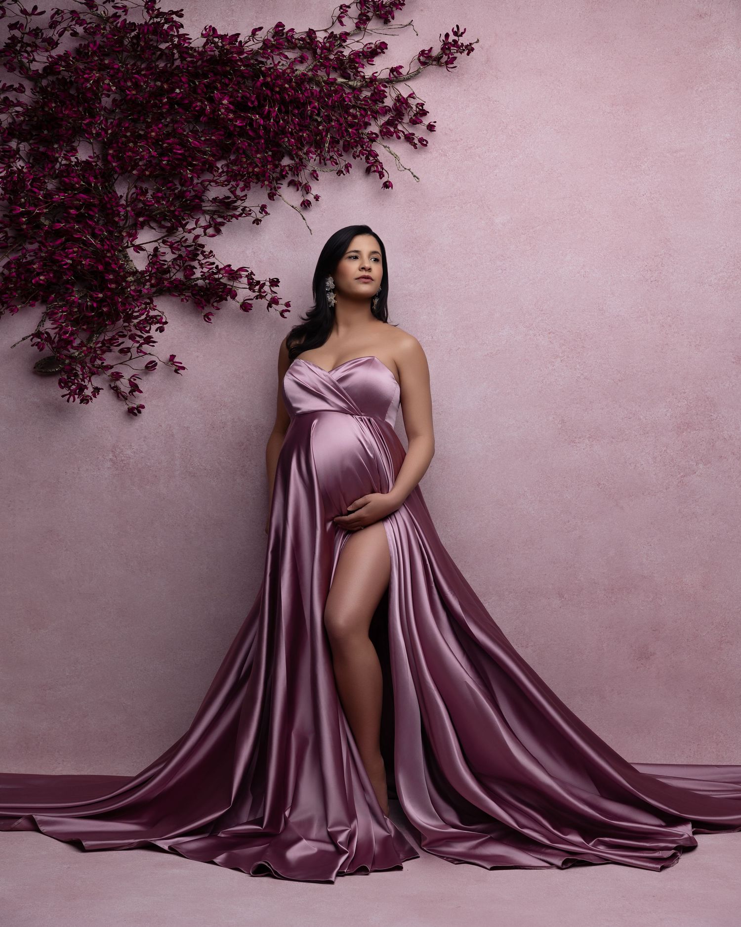 A maternity photo shoot featuring a flowing mauve silk gown against a pink wall with deep burgundy foliage.