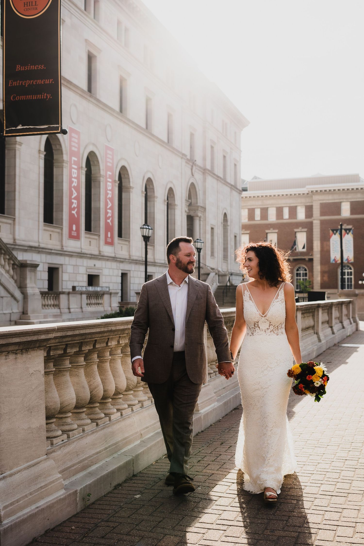 Newlyweds walk hand in hand along stone balustrade of historic building.