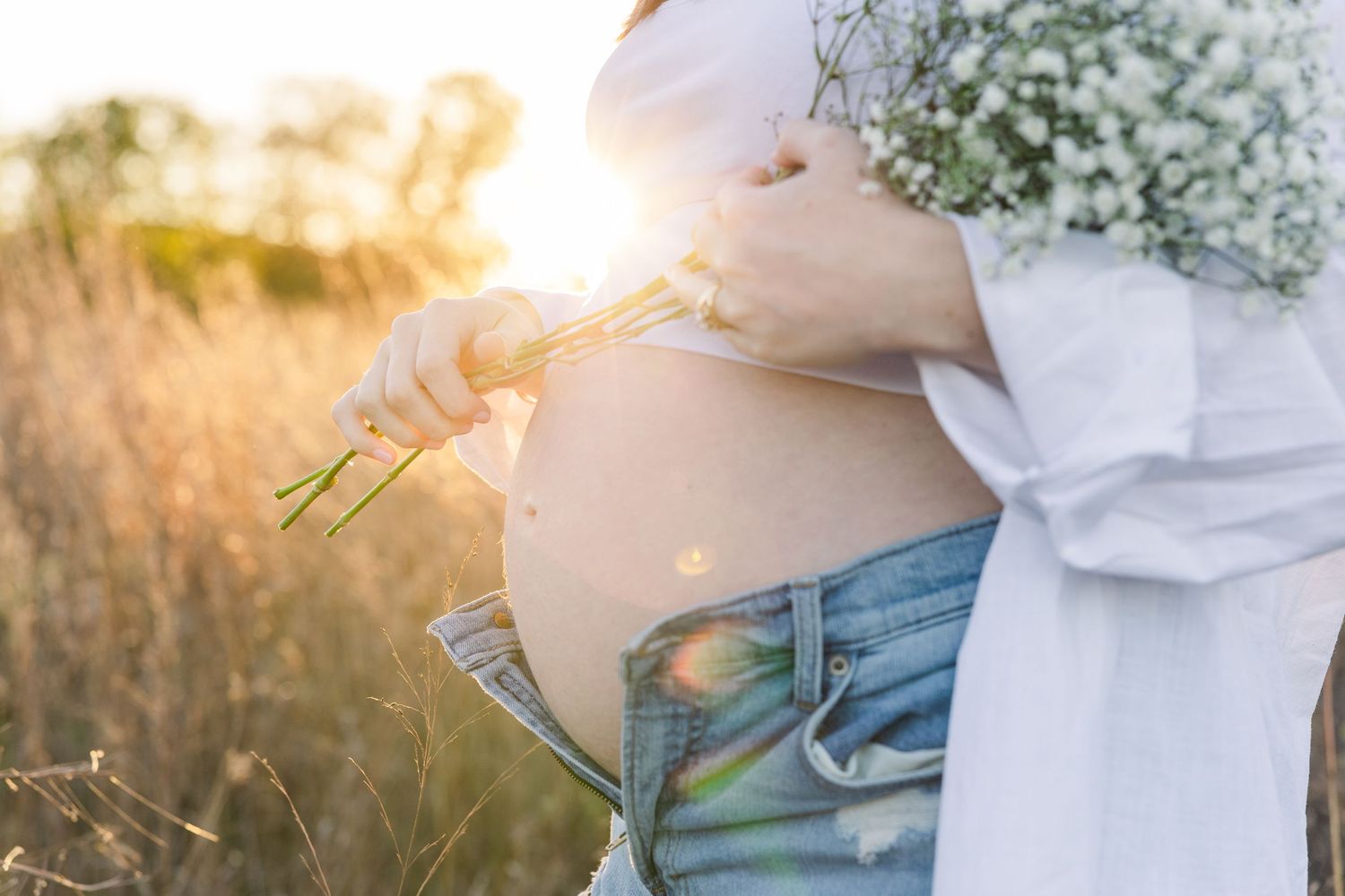 Close-up of pregnant belly with baby's breath flowers at sunset.