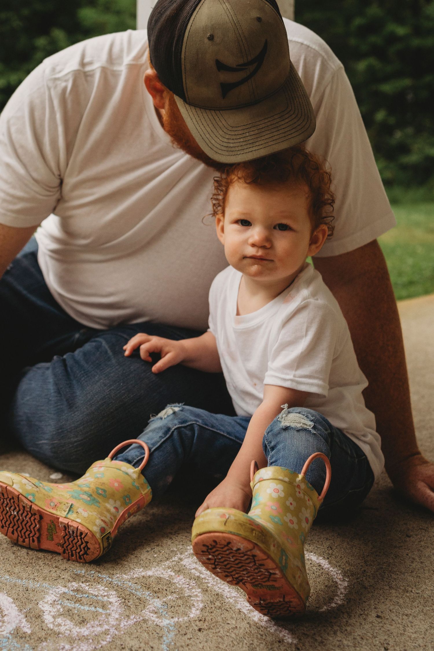 A toddler in colorful rain boots sits on concrete with adult beside them.