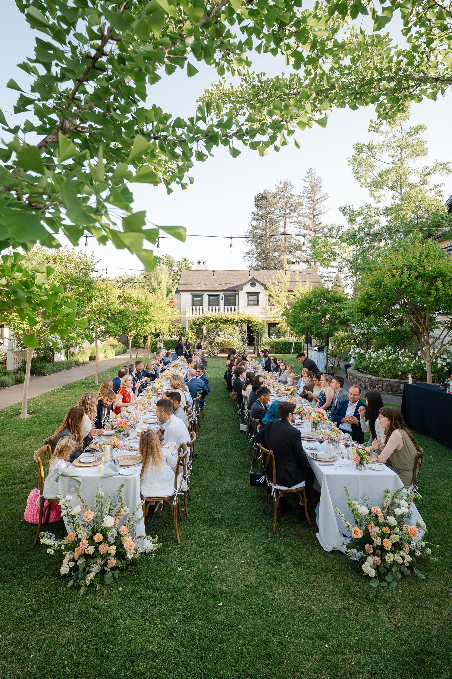 Elegant outdoor wedding reception with long tables, floral centerpieces, and guests dining on a lush green lawn.