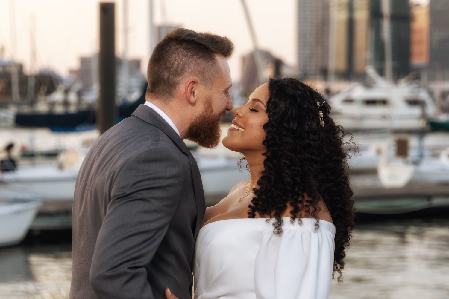 Romantic couple sharing an intimate moment at a marina waterfront, woman in white off-shoulder dress with curly hair.