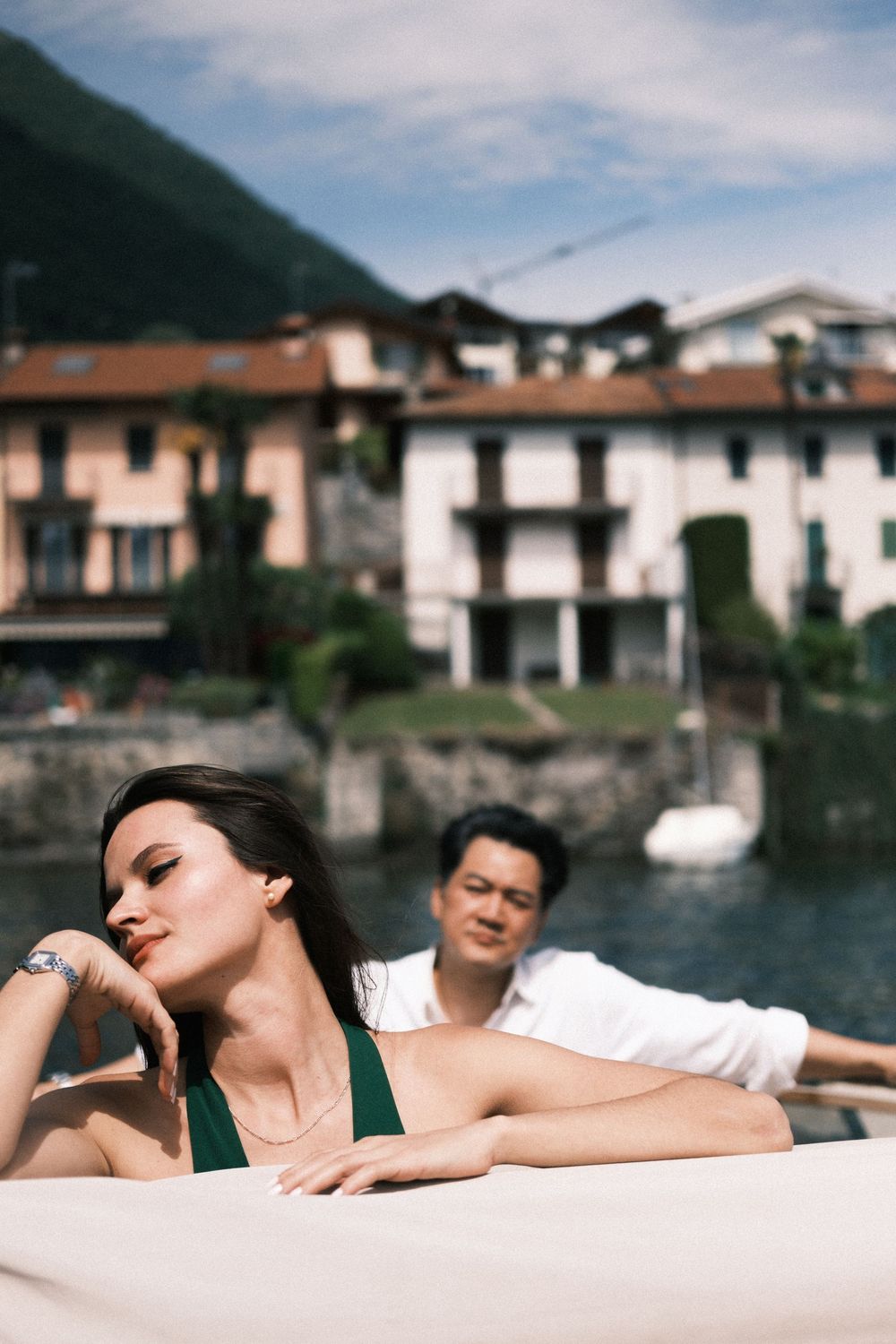 A woman in a green swimsuit relaxes on a boat with a man in white shirt, scenic Italian lakeside village in background.