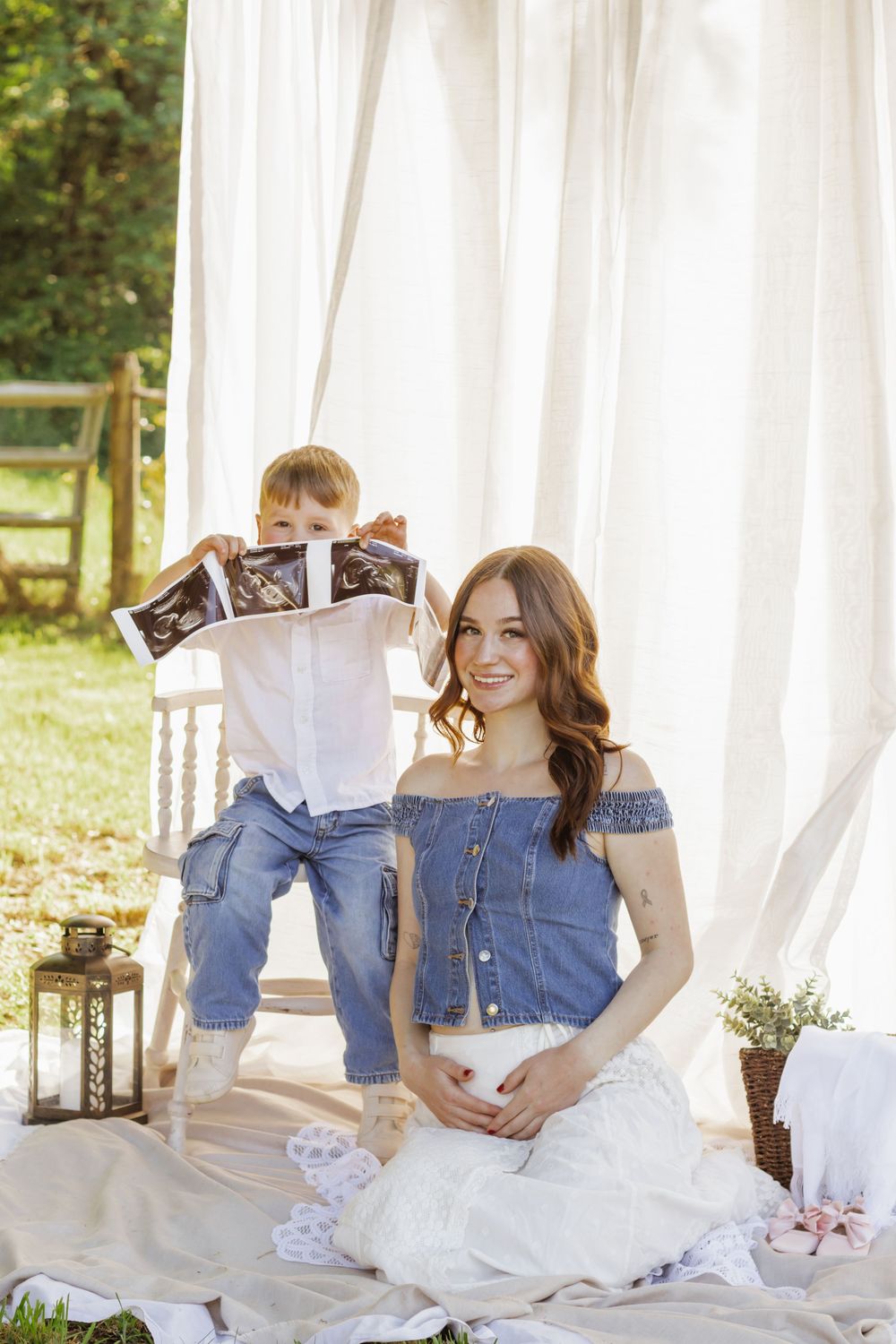 pregnant woman photographed in denim top and white skirt sitting outdoors with young boy holding ultrasound photo by roy wa photographer.