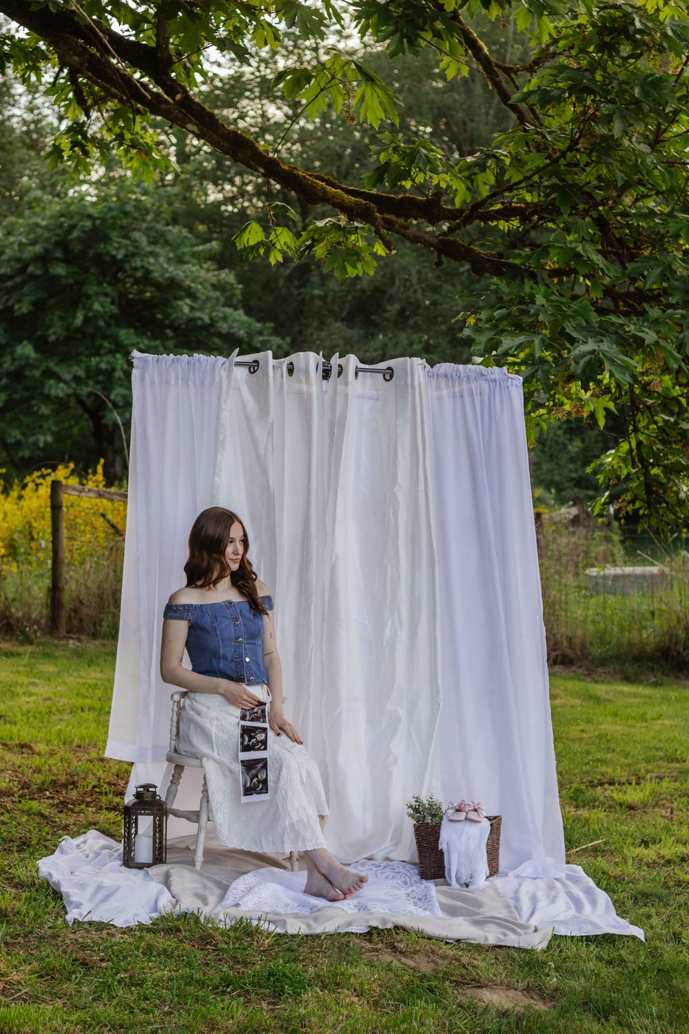 Pregnant woman photographed in denim top and white skirt sitting on chair with outdoor white curtain backdrop by Roy WA photographer