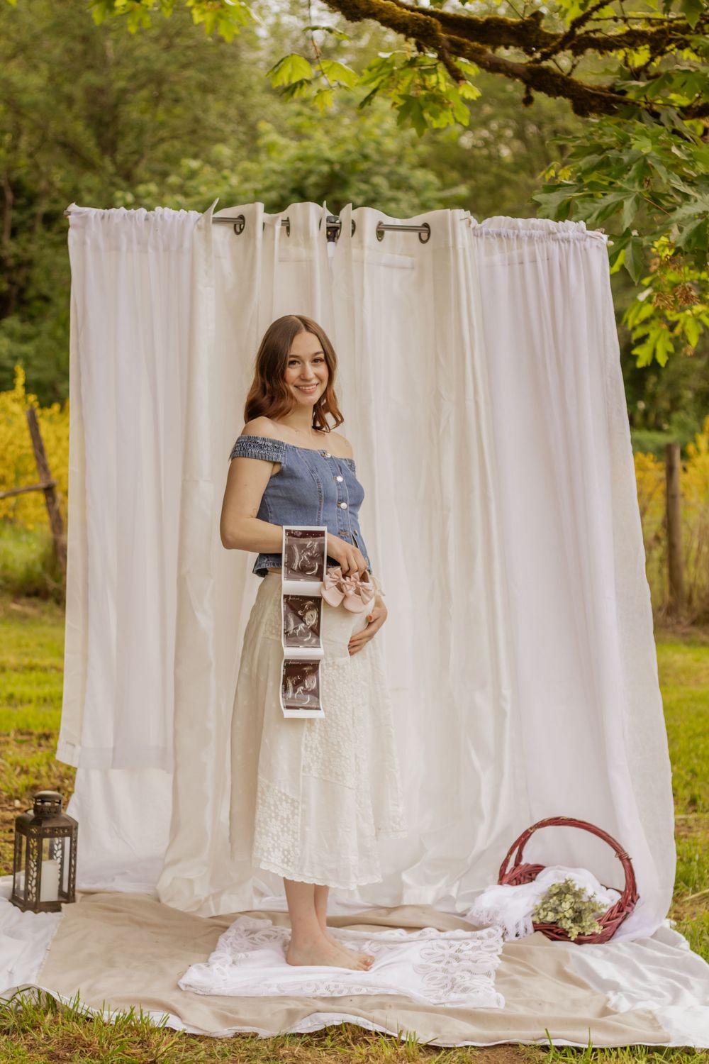 Pregnant woman in denim top and white skirt holding ultrasound photos outdoors with white curtain backdrop by pierce county photographer