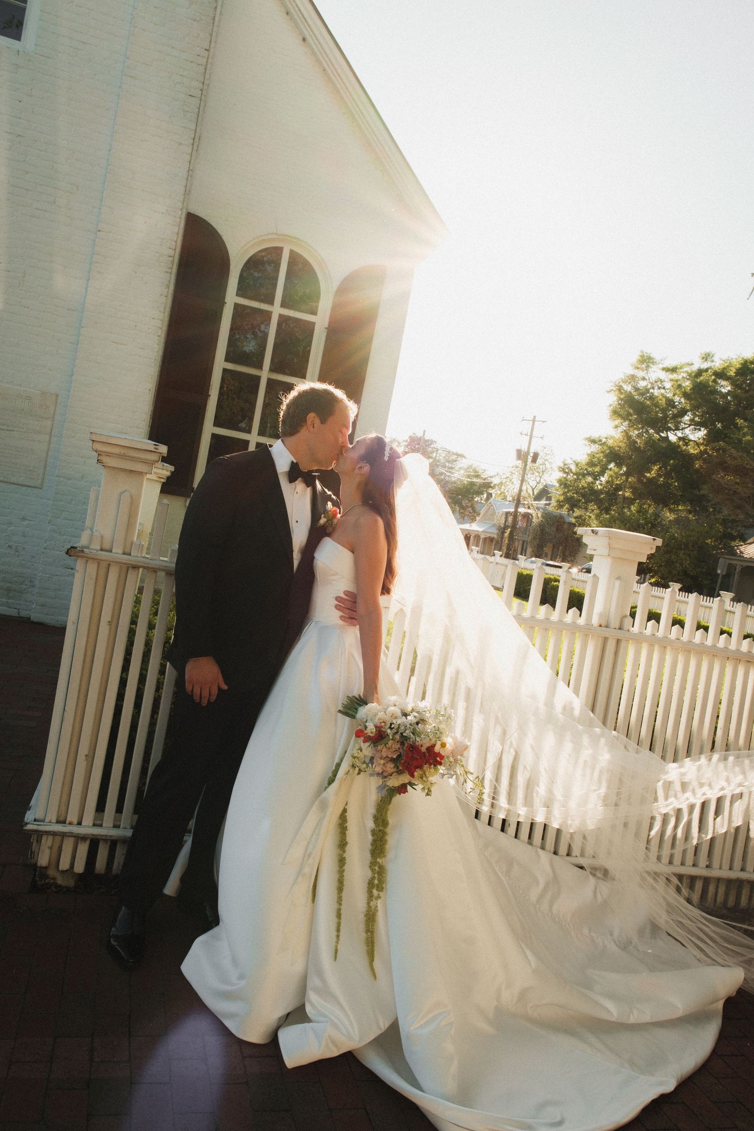 Bride and groom pose together outdoors near a white fence and chapel, bride holding bouquet, sunlight glowing behind them.