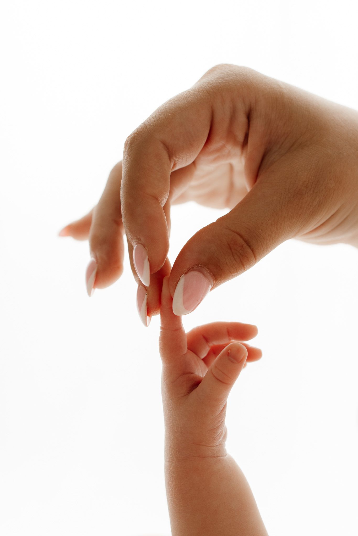 A mother's hand with French manicure gently touching her newborn baby's tiny fingers on white background.