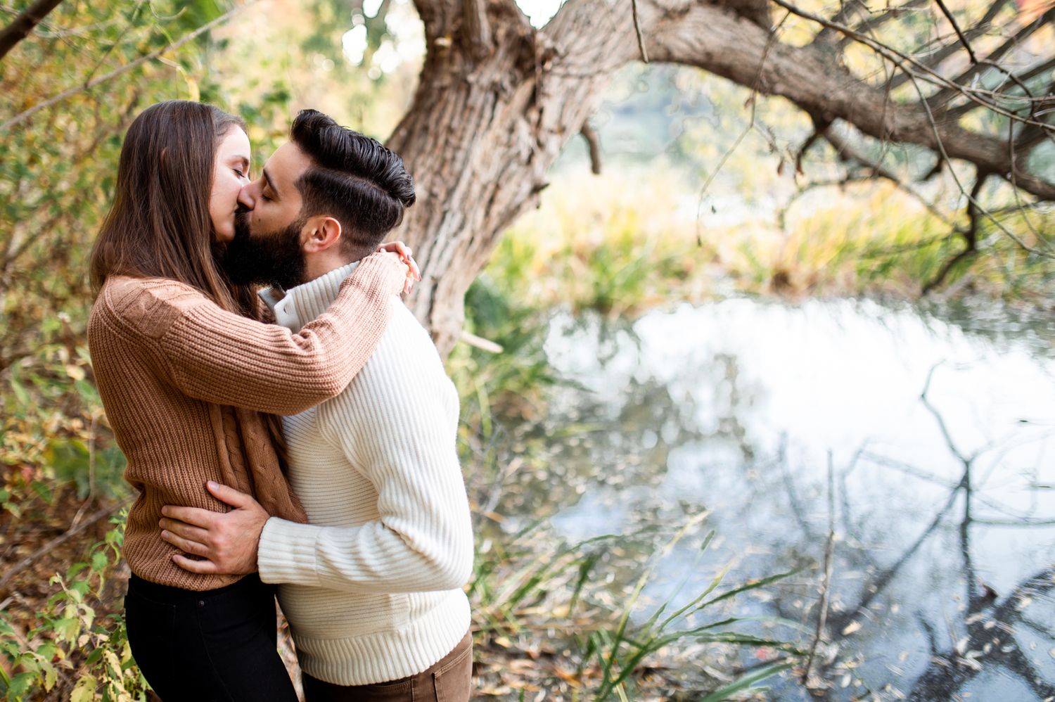 A couple shares a romantic kiss near a serene pond surrounded by autumn trees in a peaceful outdoor setting.