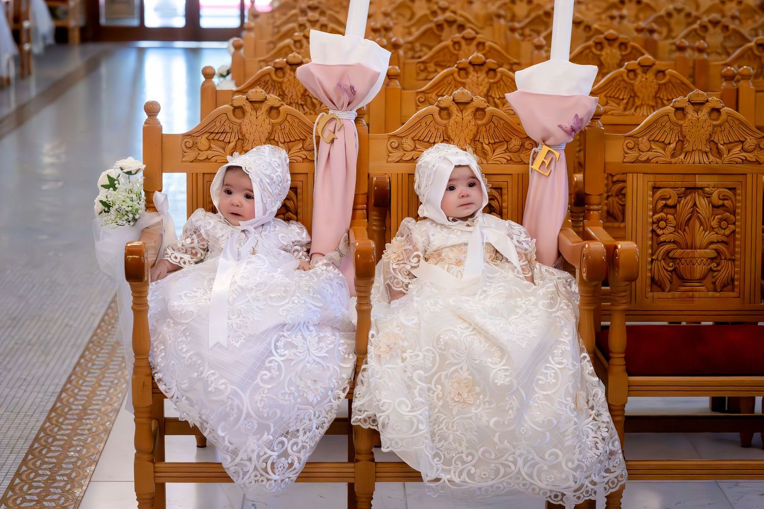 Two babies in white lace christening gowns and bonnets sit in wooden chairs holding candles during a baptism ceremony.