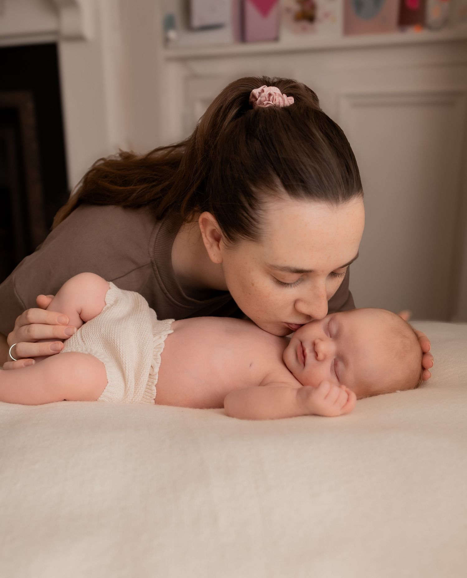 A tender mother gently kisses her sleeping newborn baby lying on a soft white blanket indoors.