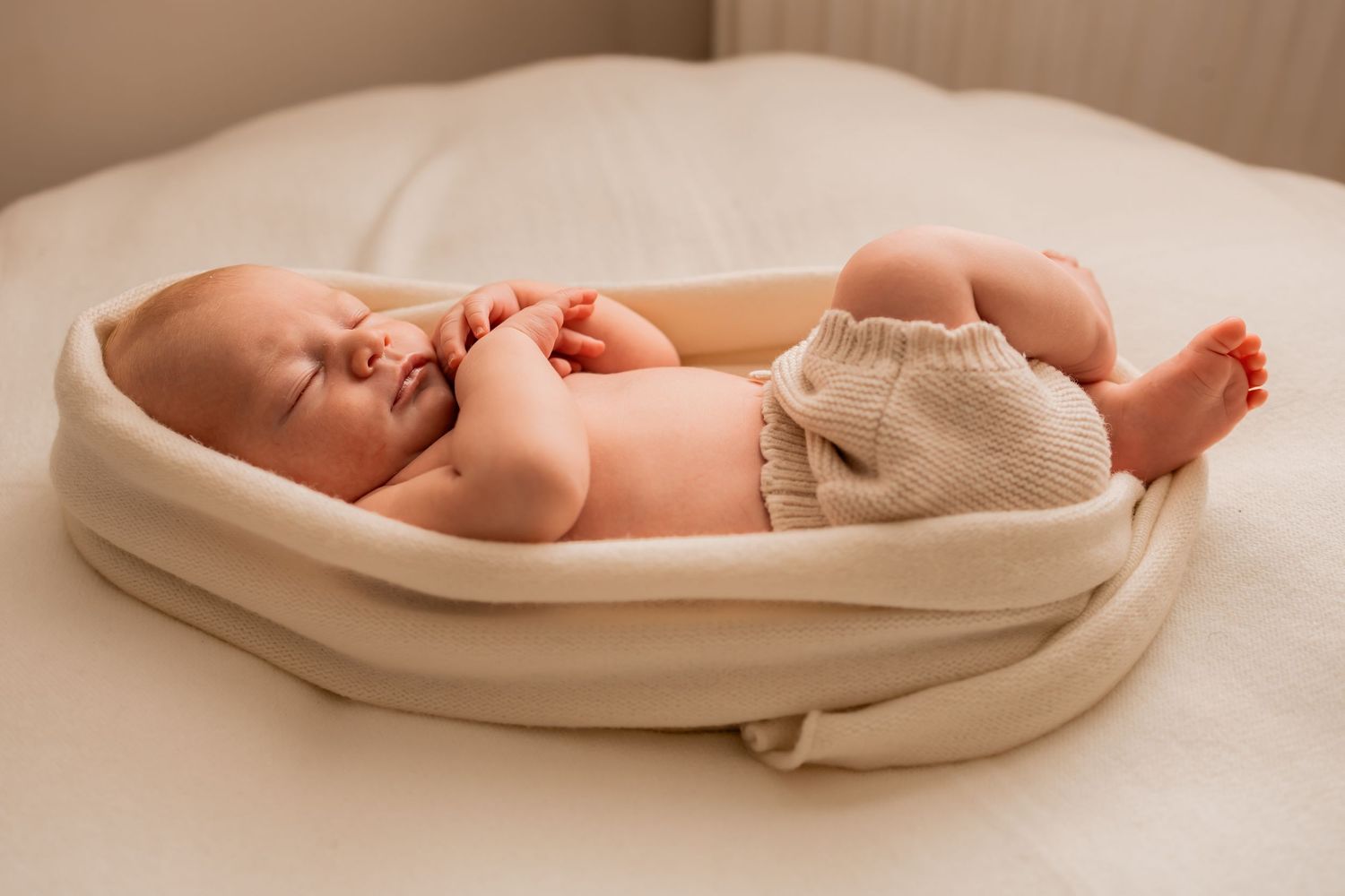 Newborn baby sleeping peacefully in a cream knit wrap on a white bed, wearing beige bloomers.