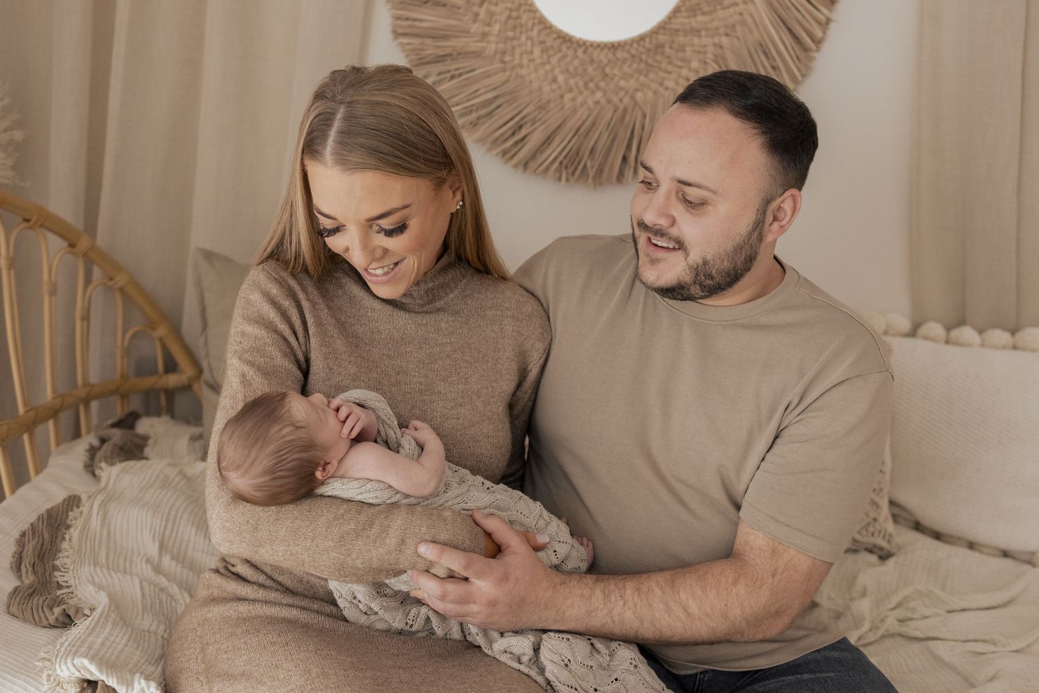 A couple sits together on a bed, lovingly cradling their newborn baby in a cozy, neutral-toned nursery setting.