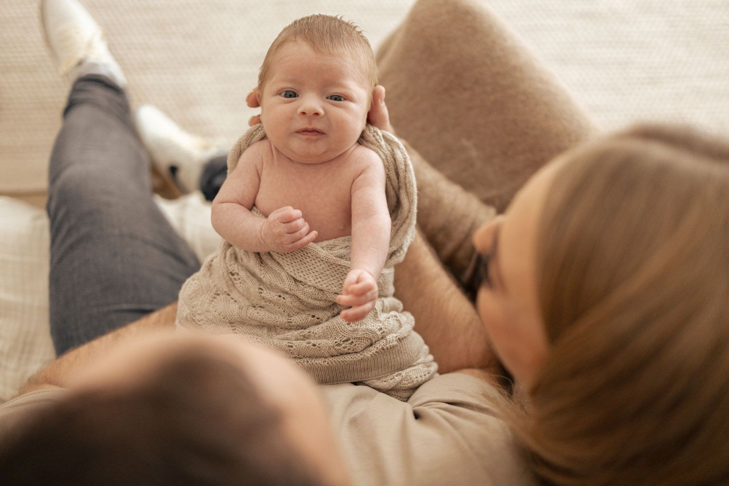 Newborn baby wrapped in a knit blanket lying on a couch with parents looking on lovingly.