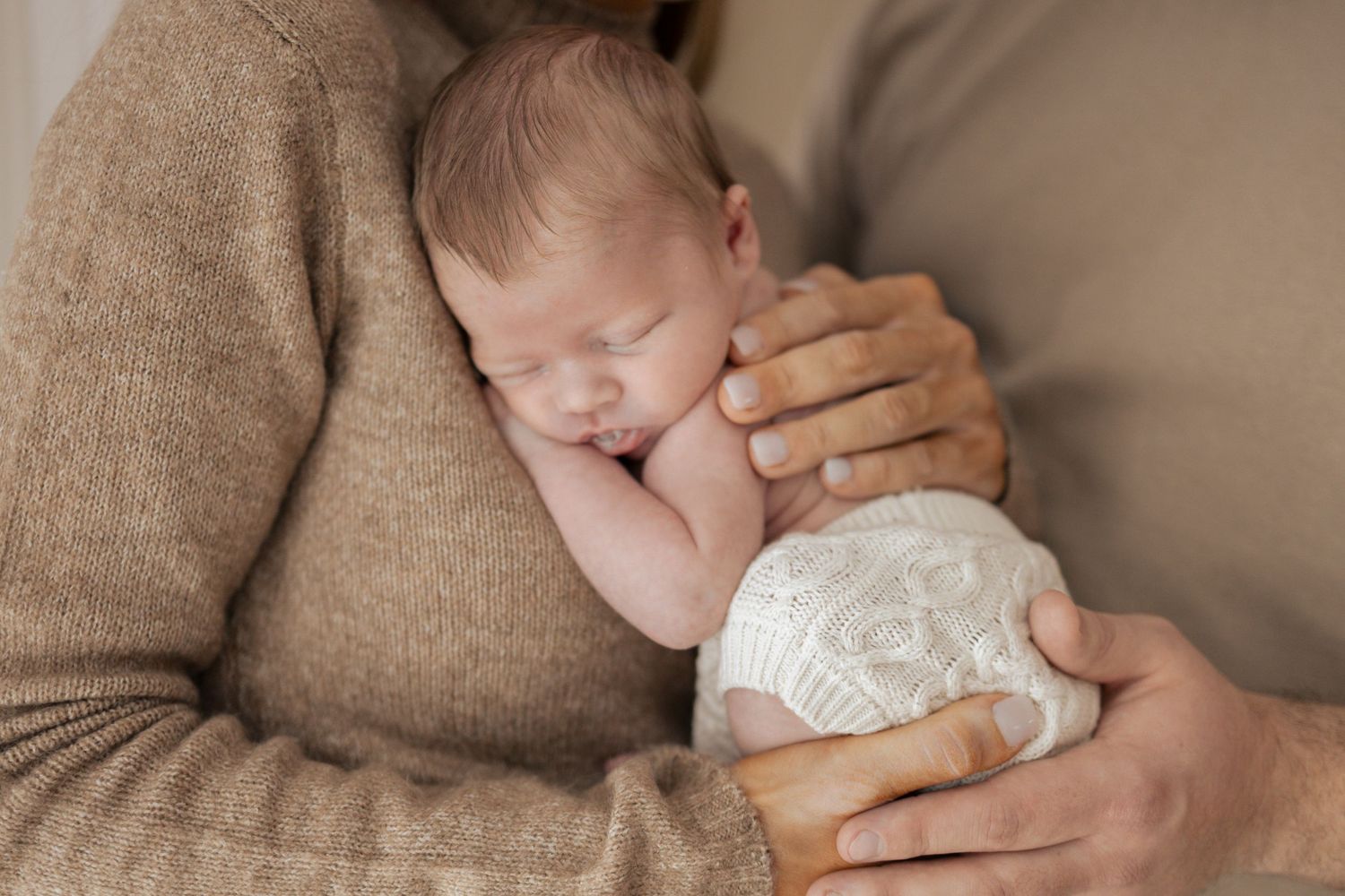 Sleeping newborn baby cradled gently in parent's arms wearing white lace outfit.