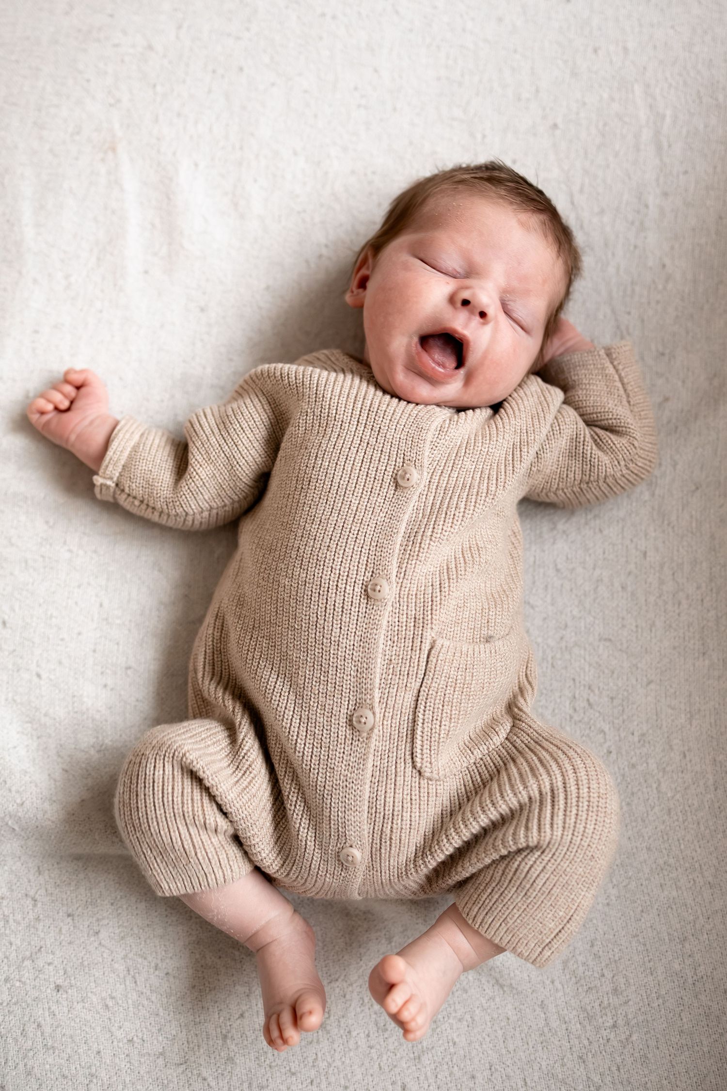 Newborn baby yawning in a beige knit romper, lying on a soft white blanket with arms stretched out.
