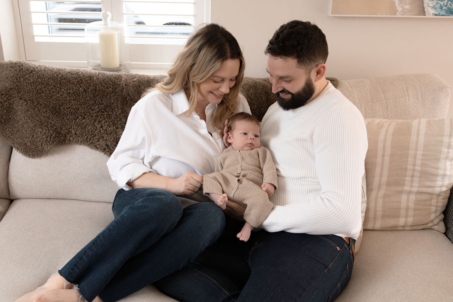 A happy couple sitting on a beige sofa, smiling and holding their newborn baby dressed in a brown outfit.