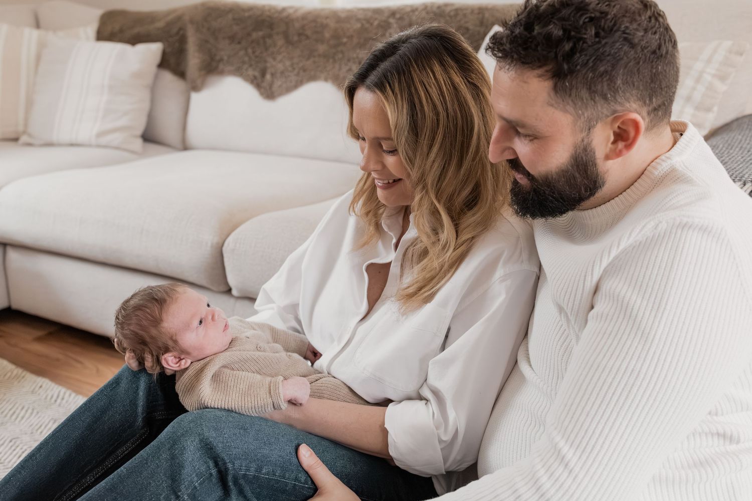 A couple sits on the floor smiling at their newborn baby in a bright, cozy living room.