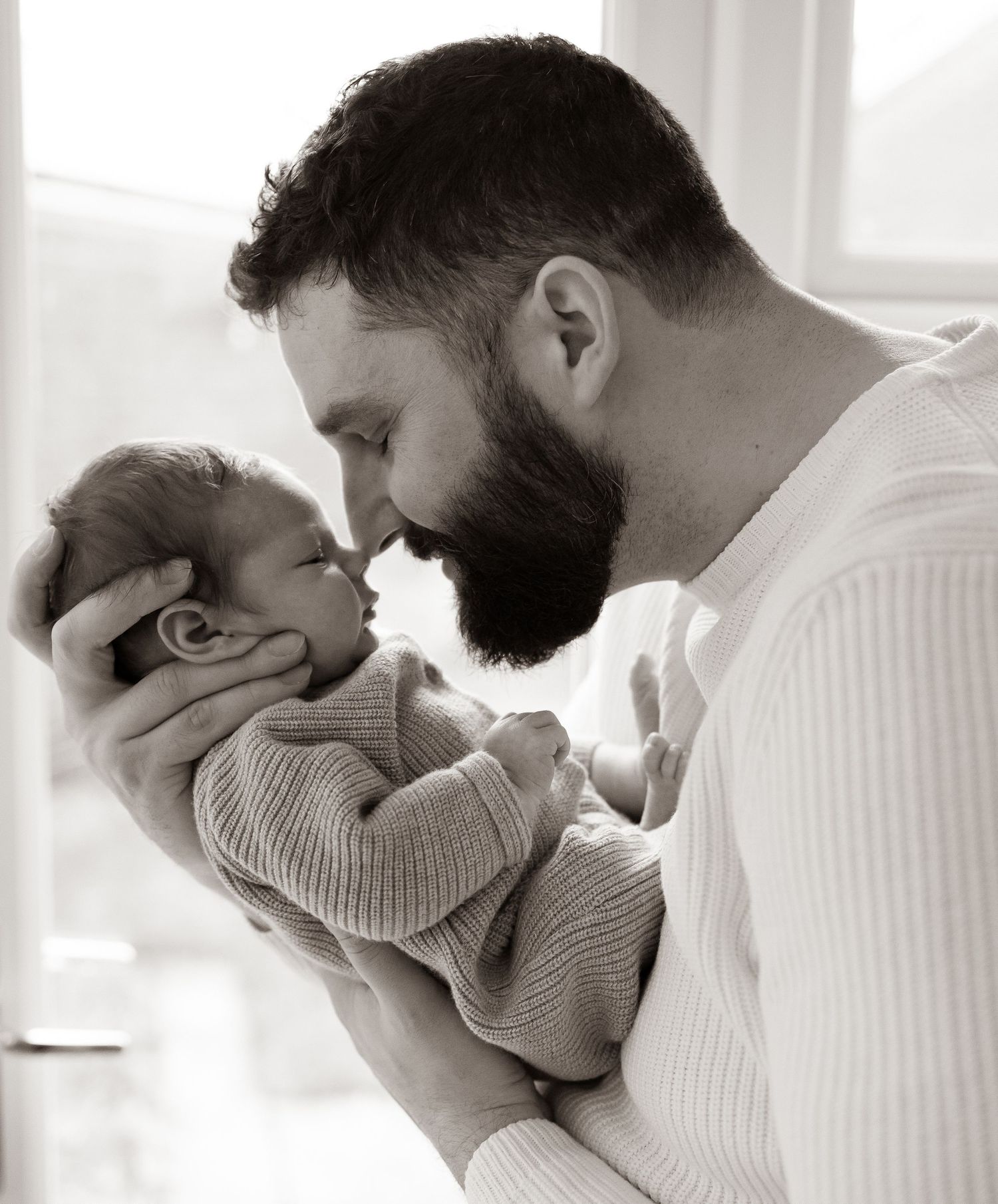 Bearded father tenderly holds newborn baby close in black and white portrait photo.