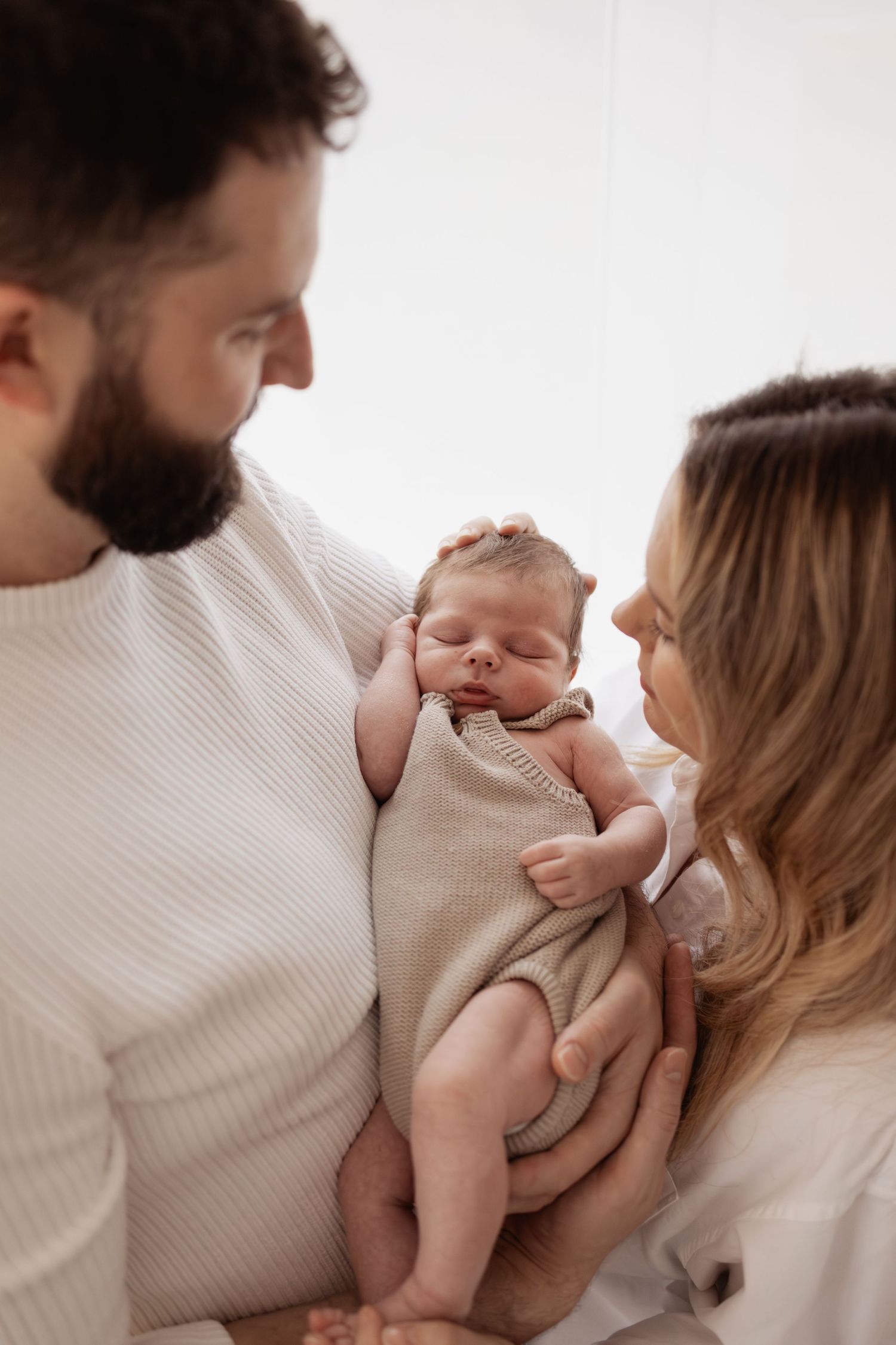 A bearded father and blonde mother tenderly hold their sleeping newborn baby wrapped in a beige knit blanket.