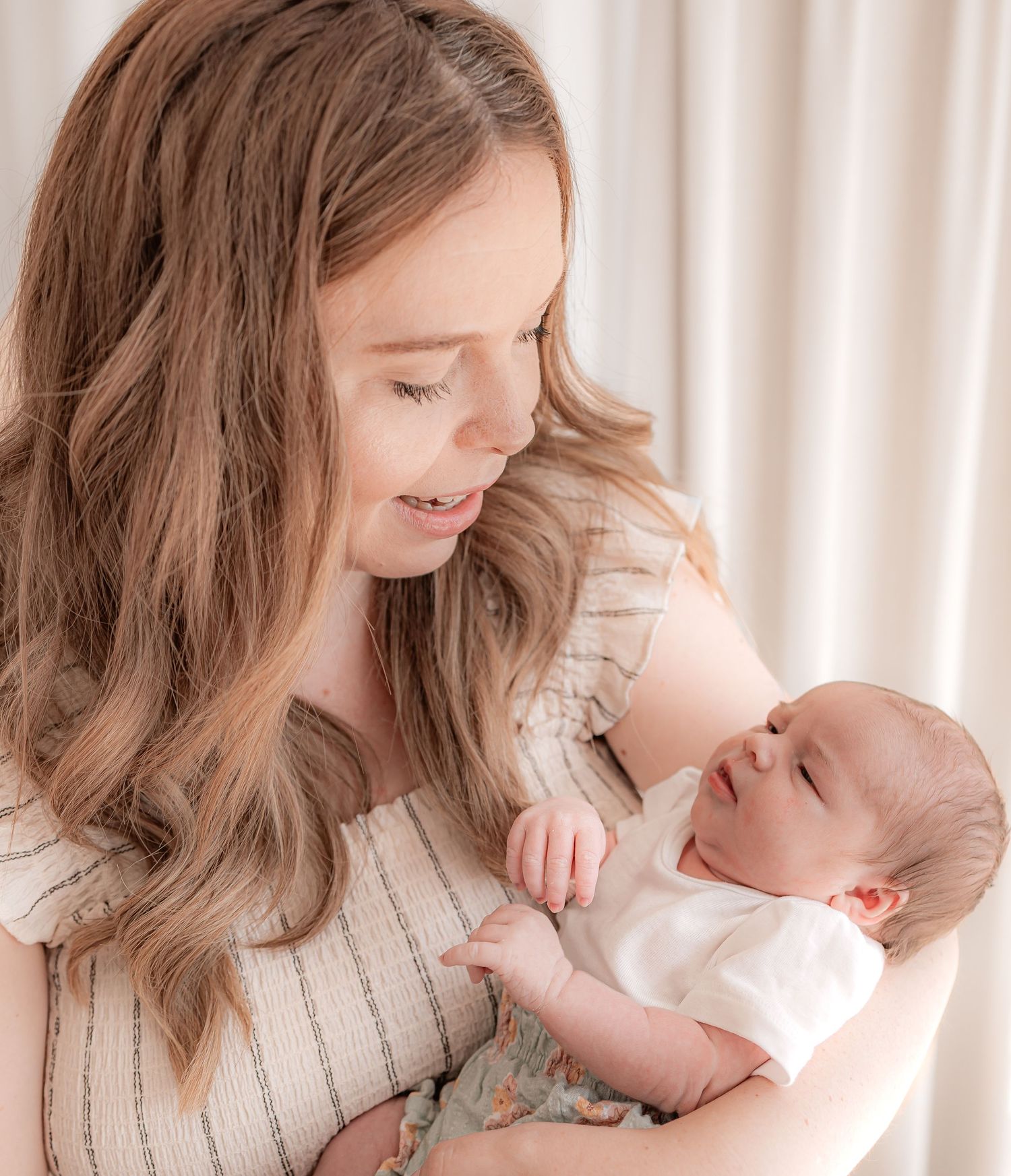 A young mother with long blonde hair tenderly holds and gazes at her newborn baby in a bright, airy studio.