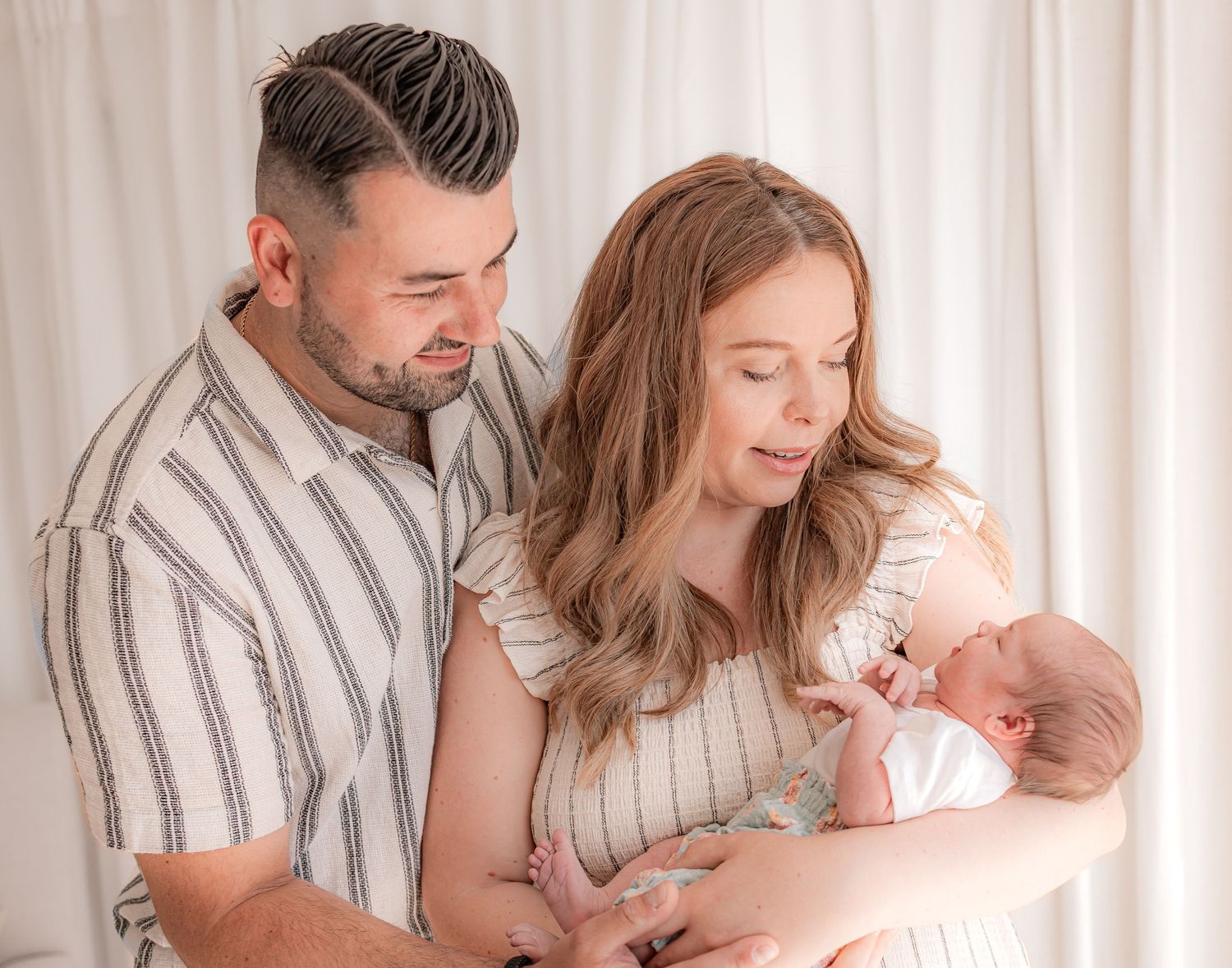 A couple tenderly gazes at their newborn baby during a professional newborn photography session with a white backdrop.