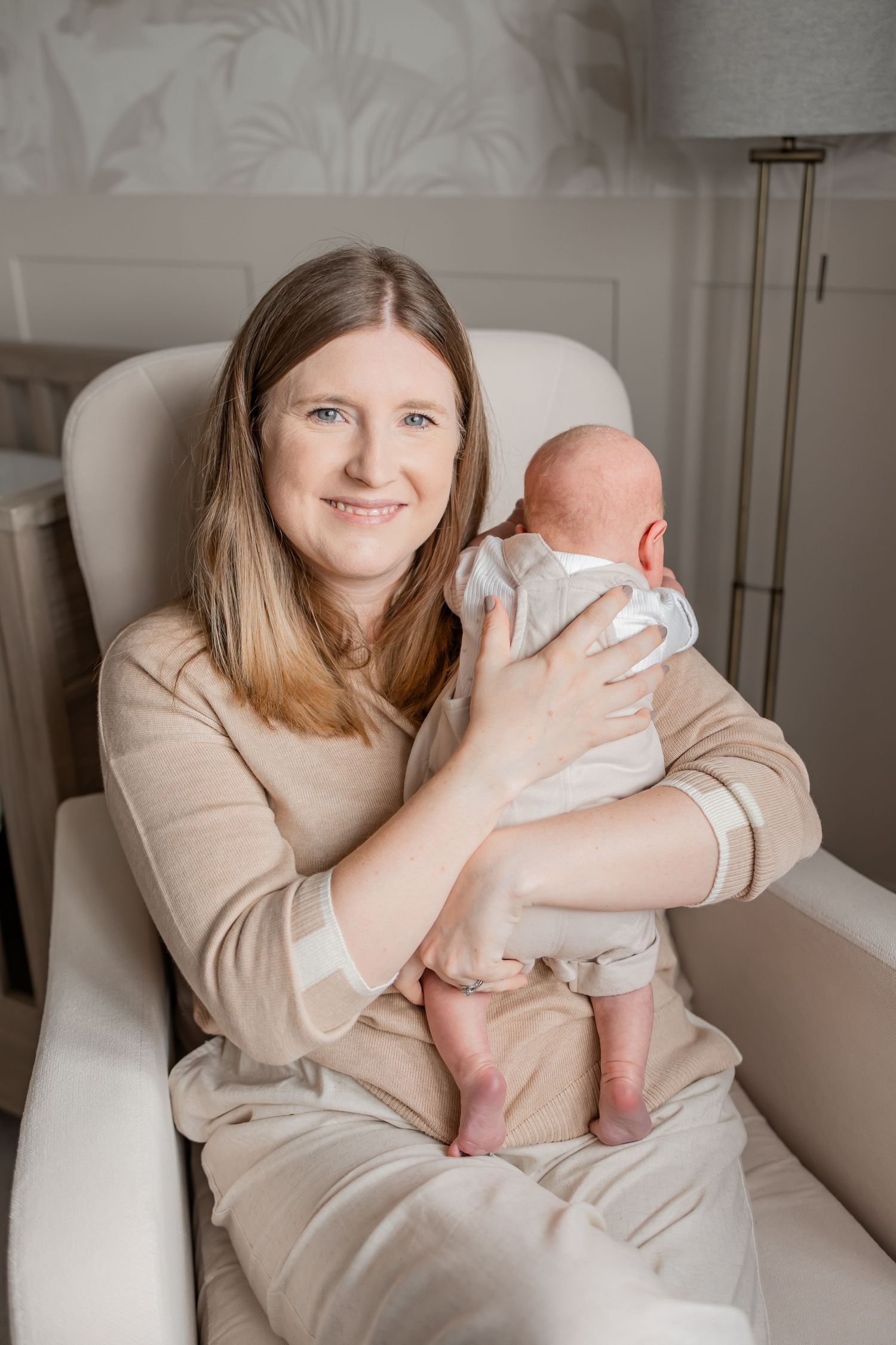 A smiling woman with long hair sits in a cream chair holding a newborn baby in a softly decorated nursery room.
