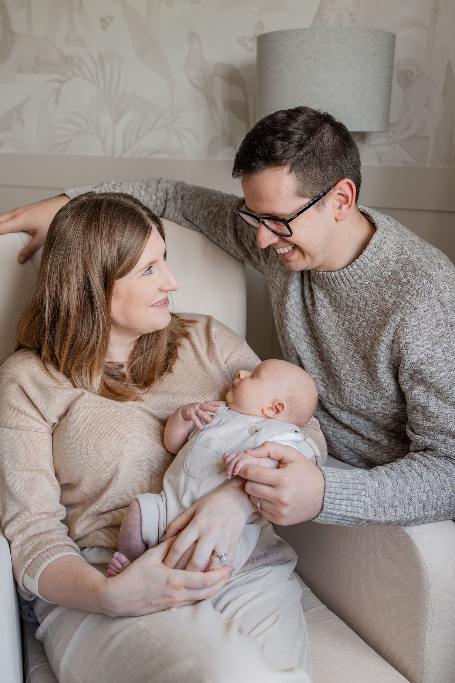 Happy parents smiling at their newborn baby while sitting together in a cozy nursery chair at home.