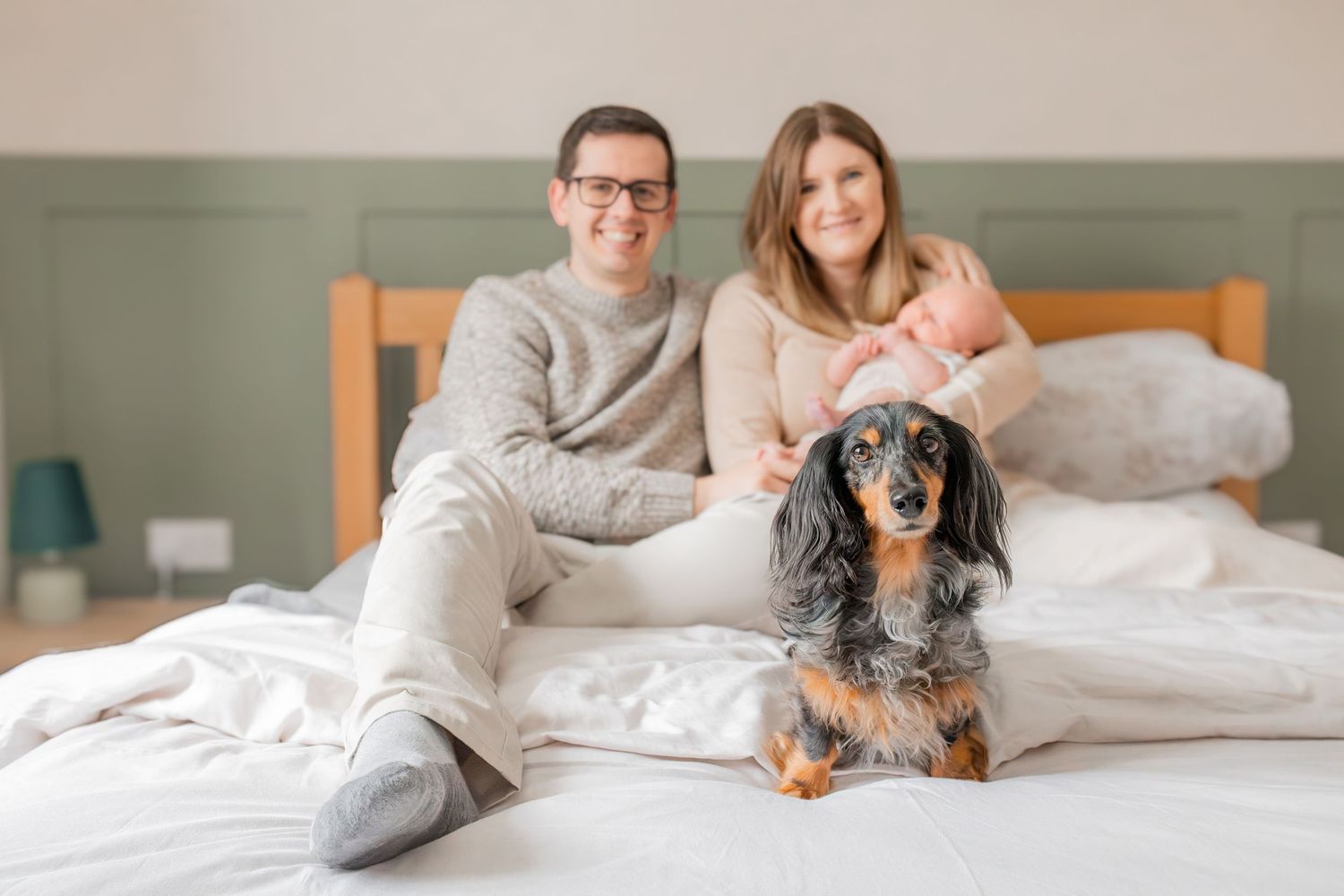 A couple sits on a white bed holding a newborn baby, with a dachshund dog in front, in a cozy bedroom.