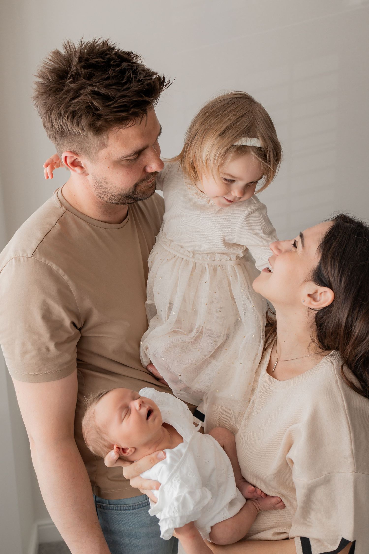 A happy family of four with a father, mother, toddler girl, and newborn baby posing together in neutral tones.