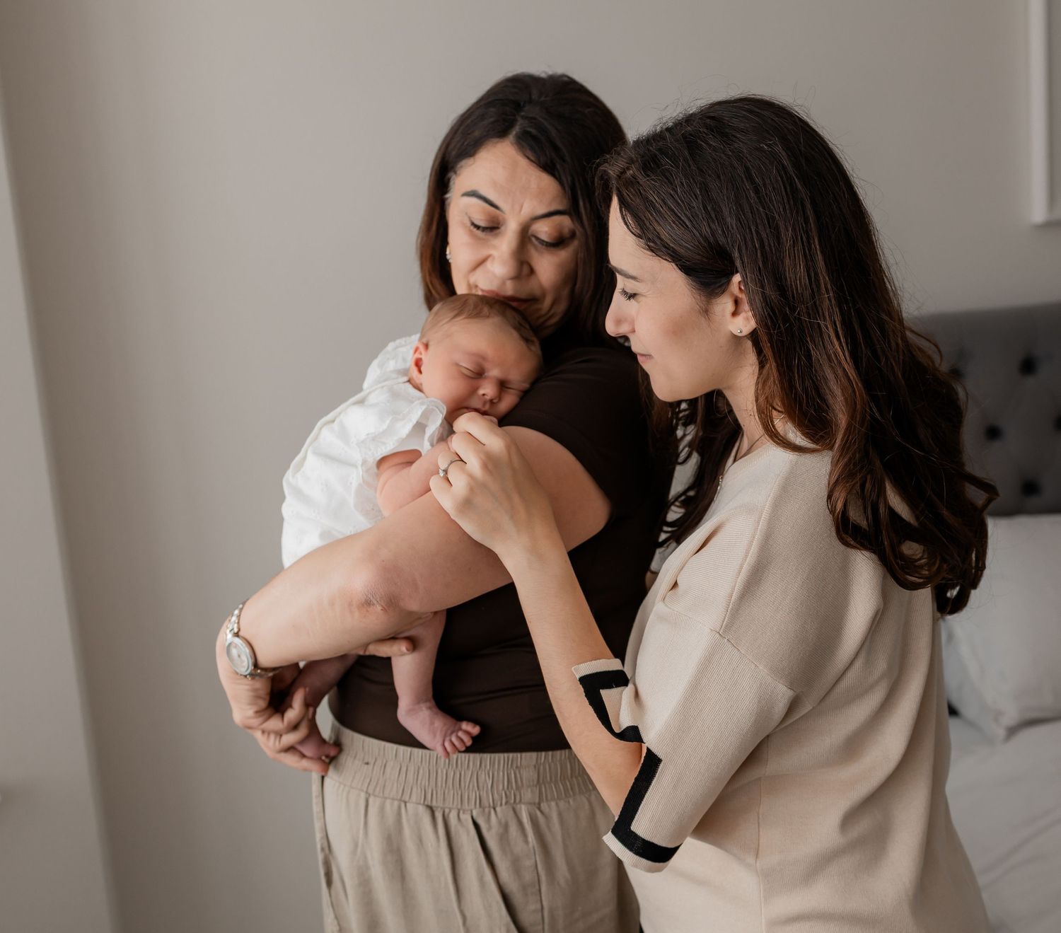 Two women admiring a newborn baby indoors, one holding the infant while the other gently touches its hand.
