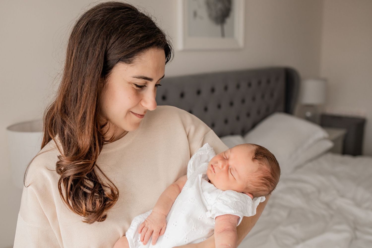 A young brunette mother tenderly holds her sleeping newborn baby in a softly lit bedroom.