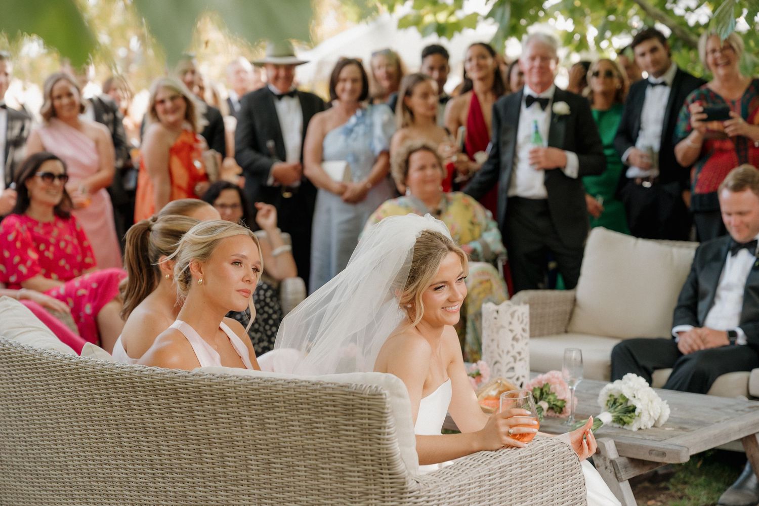 Bride and bridesmaid seated outdoors at elegant wedding ceremony with guests gathered in background.