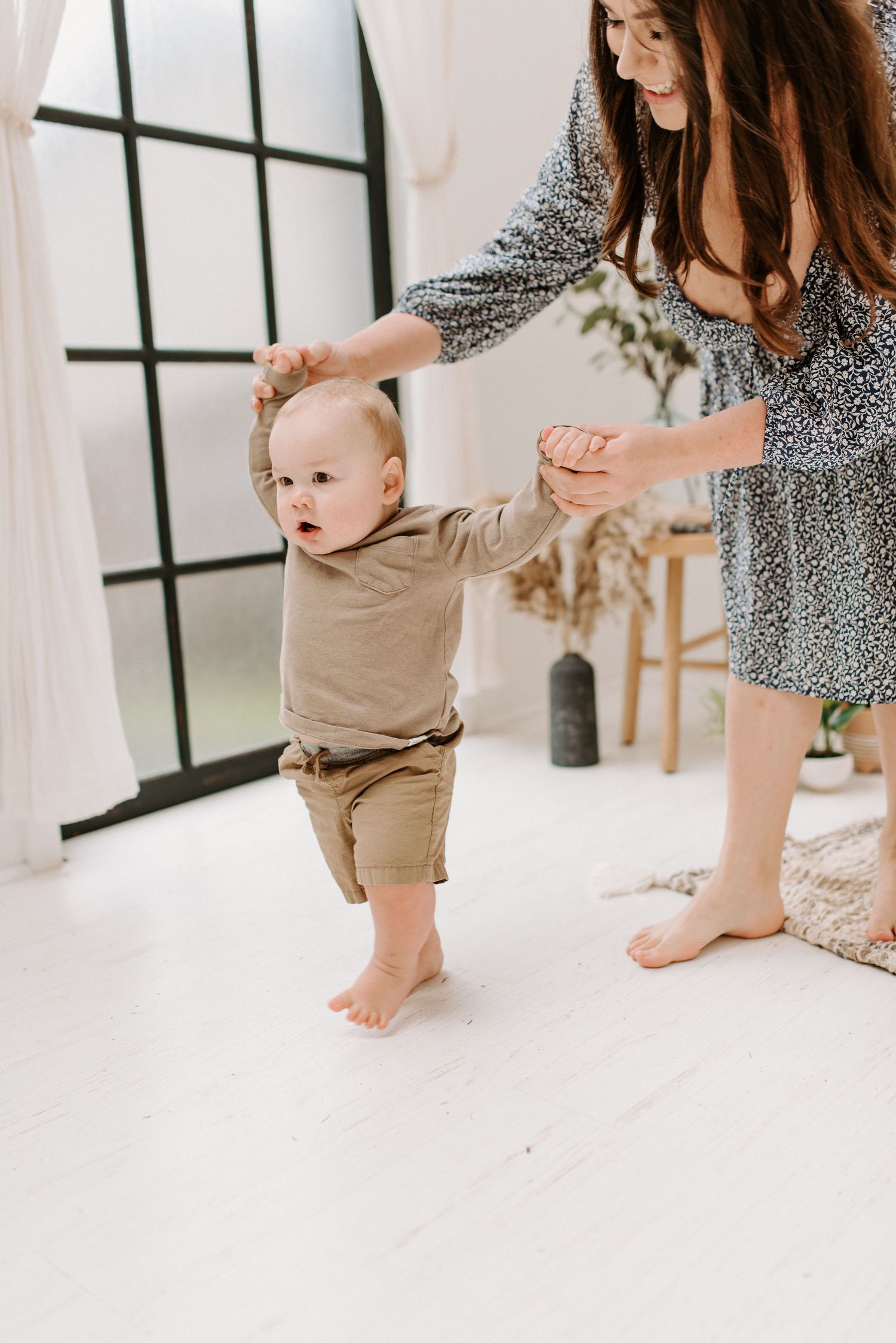 A mother helps her baby take first steps on a white floor in a bright, modern room.